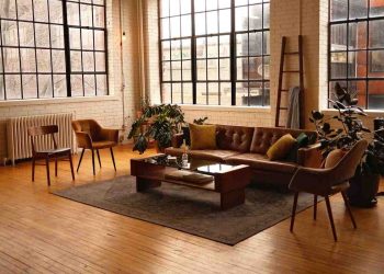 Dnevna soba sa prozorima u industrijskom stilu, kožnim kaučem svetlo braon boje, parketom, biljkama i merdevinama naslonjenim uza zid