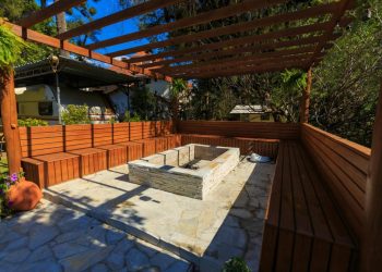 Drvena pergola sa ugrađenim klupama i pravougaonim kamenim ognjištem.