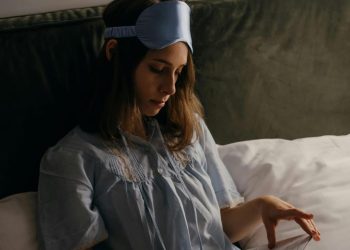 Osoba u plavoj spavaćoj košulji sa maskom za spavanje na čelu sedi na krevetu i gleda u knjigu.