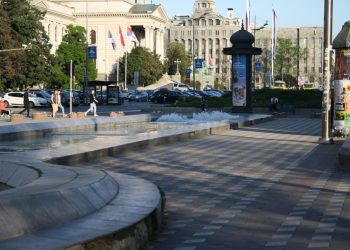 Trg sa plitkim fontanama i pešacima koji prolaze pored njih.