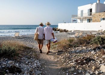 Dve osobe u šeširima nose torbu i peškire dok idu ka kamenitoj plaži