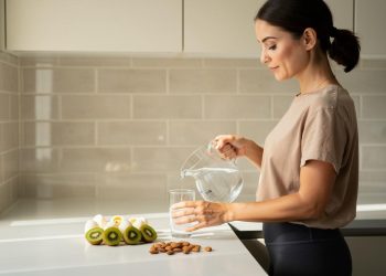 Žena u kuhinji sipa vodu iz bokala u čašu pored presečenih kivija i badema