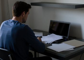 Muškarac sedi za radnim stolom pored otvorenog laptopa i razbacanih papira