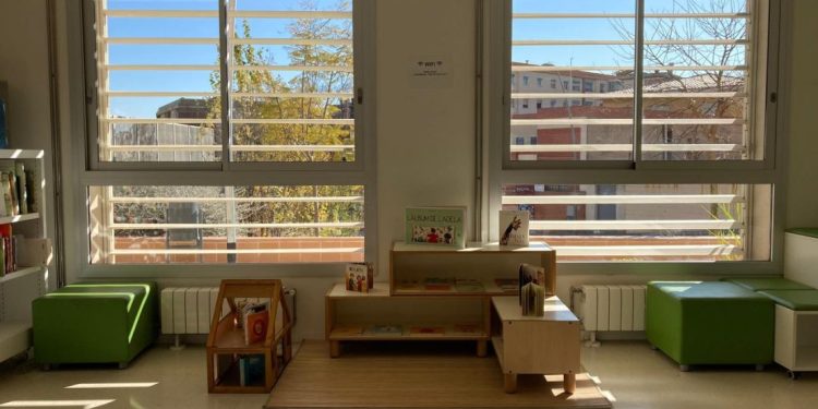 Niska polica sa dečjim knjigama ispred dva velika prozora sa žaluzinama
